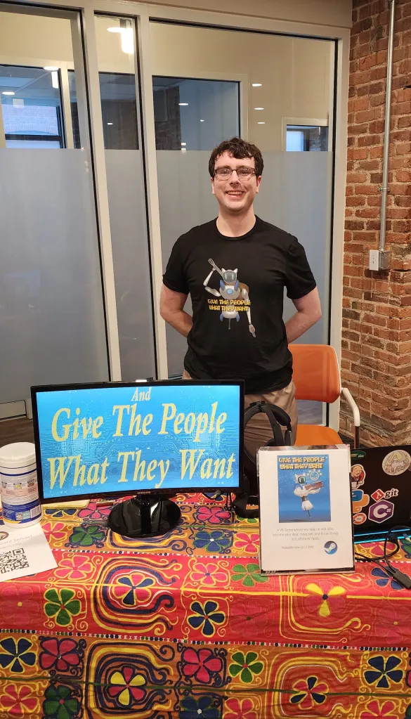 Person at a game showcase with the logo of Give the People What They Want on a monitor, VR equipment for the game in view on the desk.